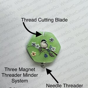 May include: A green thread cutter with a bird and floral design. The text "Thread Cutting Blade" is above the cutter. The text "Three Magnet Threader Minder System" is below the cutter. The text "(Built in magnet and two loose magnets)" is below the text "Three Magnet Threader Minder System". The text "Needle Threader" is below the cutter on the right side.