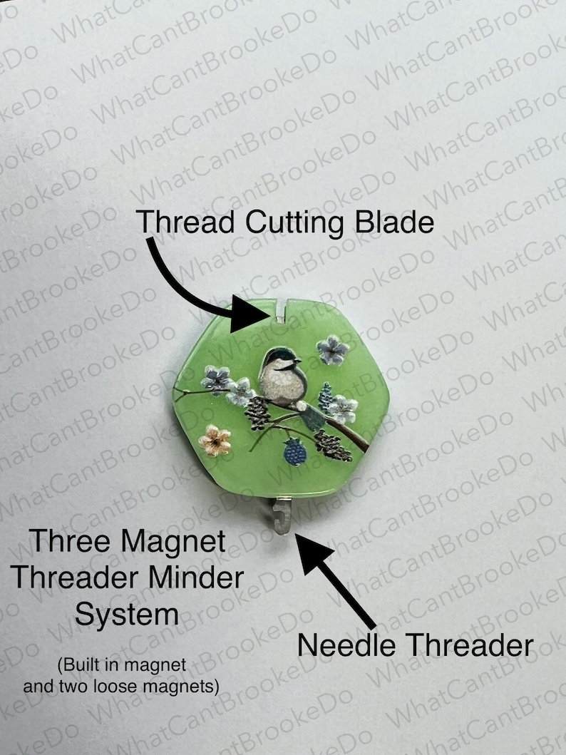 May include: A green thread cutter with a bird and floral design. The text "Thread Cutting Blade" is above the cutter. The text "Three Magnet Threader Minder System" is below the cutter. The text "(Built in magnet and two loose magnets)" is below the text "Three Magnet Threader Minder System". The text "Needle Threader" is below the cutter on the right side.