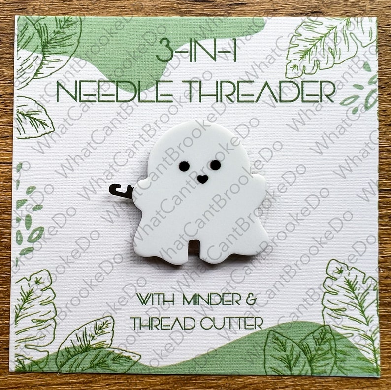 May include: White ghost-shaped needle threader with black eyes and a heart-shaped mouth. The threader has a small hook on the side. The text "3-IN-1 NEEDLE THREADER" is printed on the card. The text "WITH MINDER & THREAD CUTTER" is printed below the ghost.