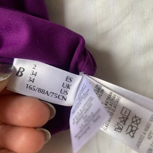 May include: A purple fabric tag with size information in black text. The tag reads "B 34 165/88A/75CN" and "ES UK US". A white tag with washing instructions is partially visible.