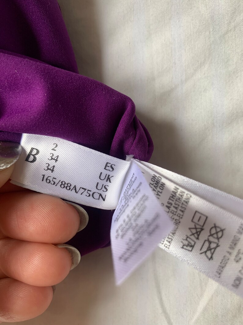 May include: A purple fabric tag with size information in black text. The tag reads "B 34 165/88A/75CN" and "ES UK US". A white tag with washing instructions is partially visible.