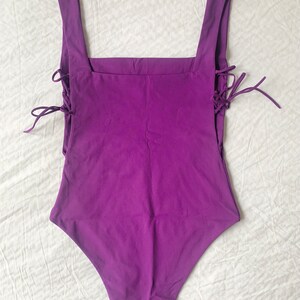 May include: A purple one-piece swimsuit with a square neckline and lace-up details on the sides.