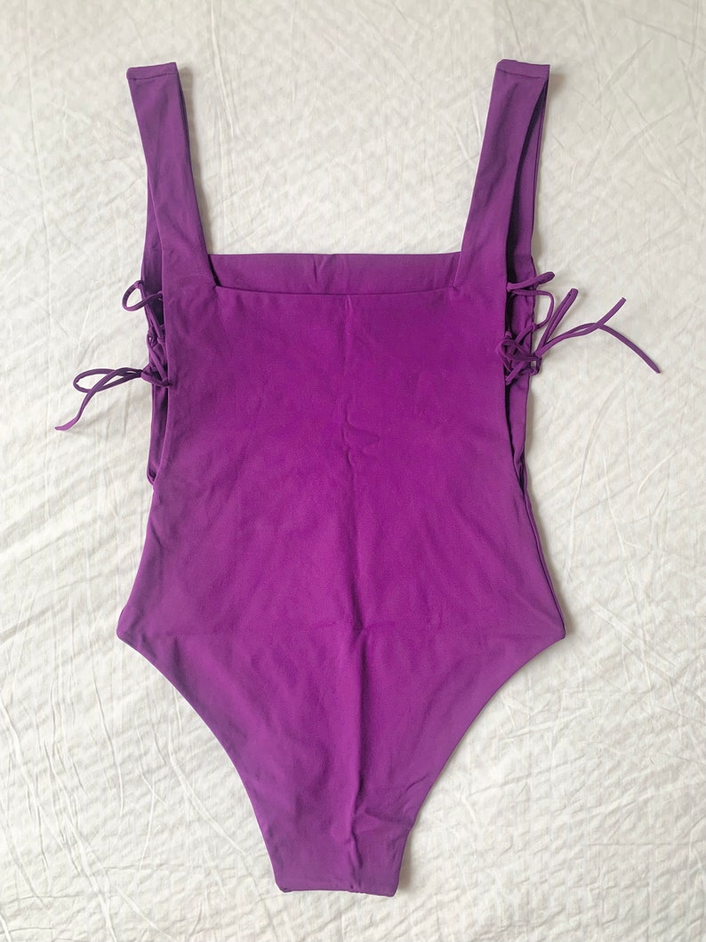 May include: A purple one-piece swimsuit with a square neckline and lace-up details on the sides.