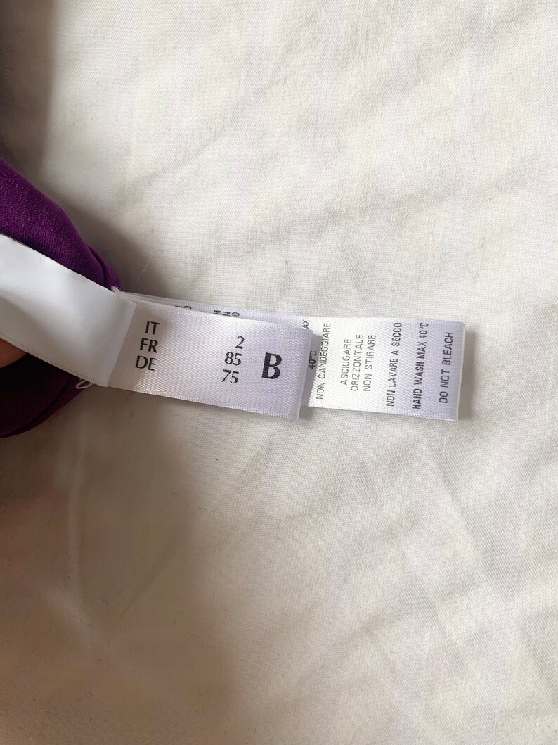 May include: A white clothing care label with black text. The label has the following information: IT, FR, DE, 2, 85, B, 75, 40&deg;C, NON CANDEGGIARE, ASCIUGARE ORIZZONTALE, NON STIRARE, NON LAVARE A SECCO, HAND WASH MAX 40&deg;C, DO NOT BLEACH.