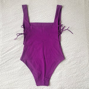 May include: A purple swimsuit with a square neckline and lace-up details on the sides.