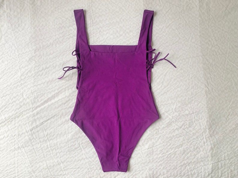 May include: A purple swimsuit with a square neckline and lace-up details on the sides.