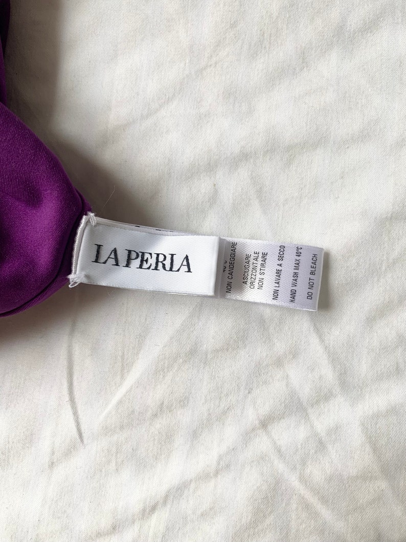 May include: A white tag with the text "LA PERIA" in black lettering. The tag has care instructions in Italian, including "NON CANDEGGIARE" (Do not bleach), "ASCIUGARE ORIZZONTALE" (Horizontal drying), "NON STIRARE" (Do not iron), "NON LAVARE A SECCO" (Do not dry clean), and "HAND WASH MAX 40&deg;C" (Hand wash at a maximum of 40 degrees Celsius).
