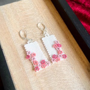 May include: A pair of rectangular drop earrings crafted from white seed beads, featuring small floral motifs in various shades of pink. Each earring is attached to a silver-toned metal leverback hook, creating a dangling design with a textured surface.