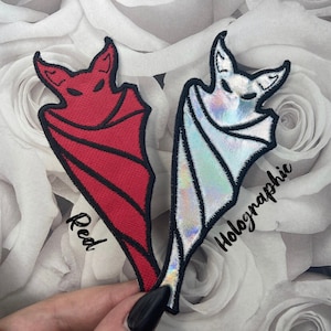 May include: Two iron-on patches of a bat in a cape. One patch is red and the other is holographic. The red patch has the word "Red" written below it. The holographic patch has the word "Holographic" written below it.