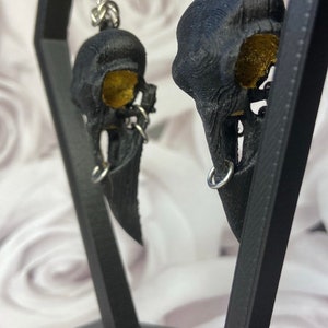 May include: A pair of black bird skull earrings with gold accents. The earrings are hanging from a black display stand.