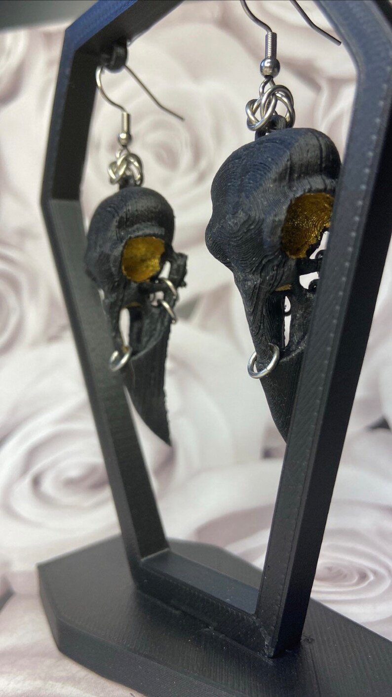 May include: A pair of black bird skull earrings with gold accents. The earrings are hanging from a black display stand.