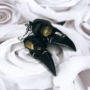 May include: A pair of black crow skull earrings with gold accents. The earrings are made of polymer clay and have a detailed design.