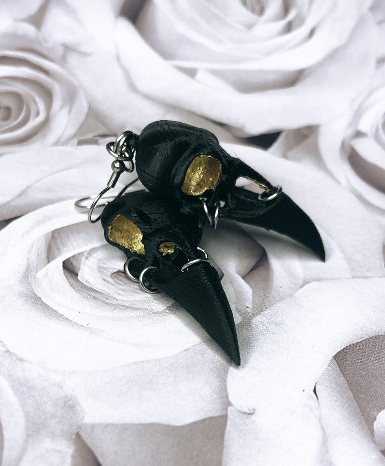 May include: A pair of black crow skull earrings with gold accents. The earrings are made of polymer clay and have a detailed design.