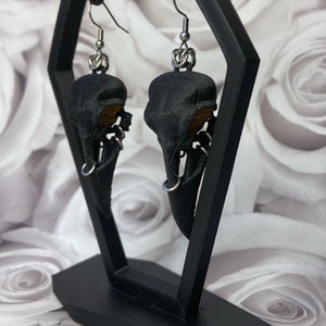 May include: A pair of black bird skull earrings with silver hooks and rings. The earrings are displayed on a black stand against a white background.