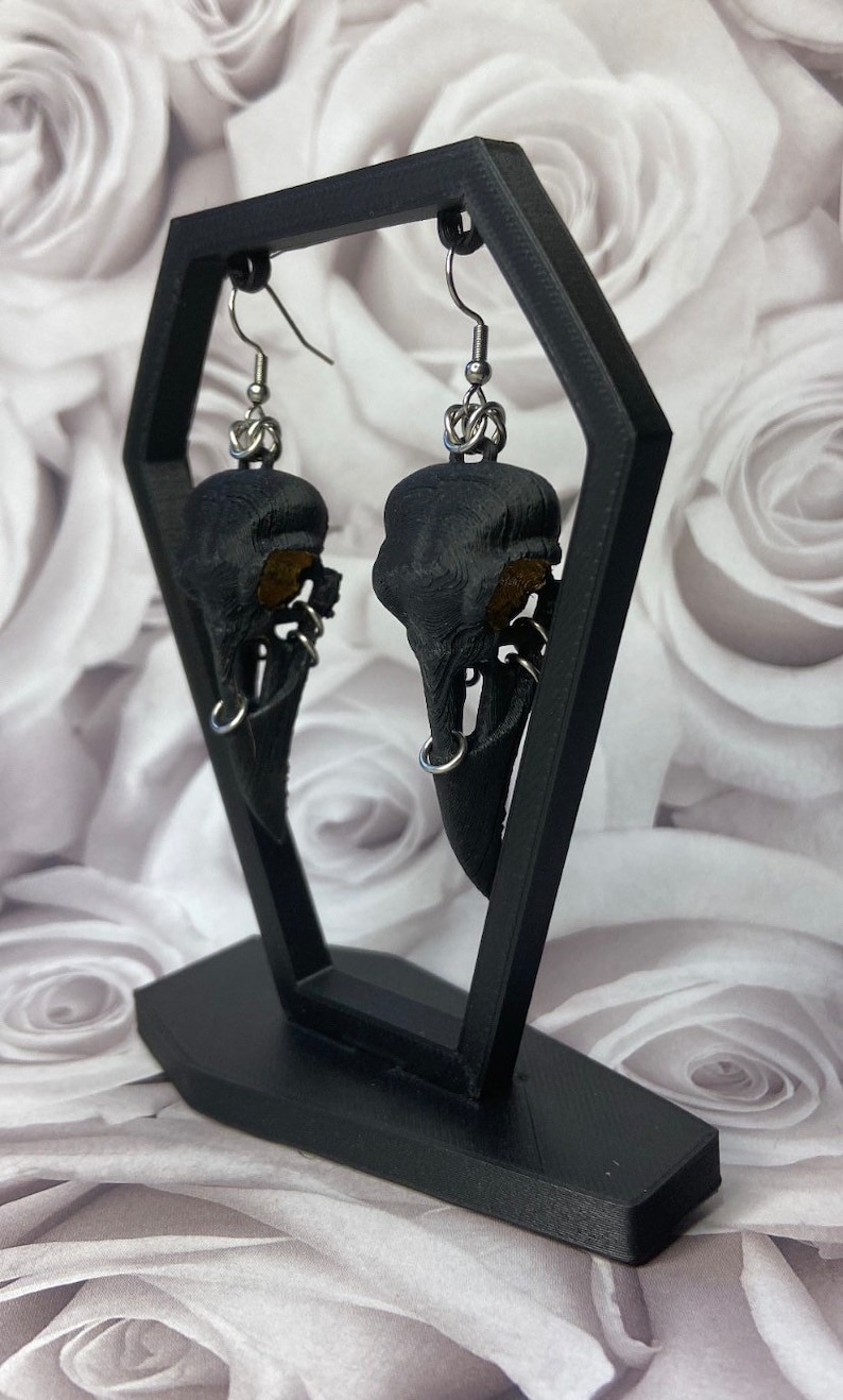 May include: A pair of black bird skull earrings with silver hooks and rings. The earrings are displayed on a black stand against a white background.
