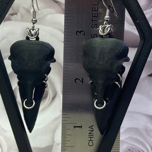 May include: A pair of black bird skull earrings with silver hoops and chains. The earrings are hanging from a ruler marked in inches and labeled "CHINA" and "STAINLESS STEEL".