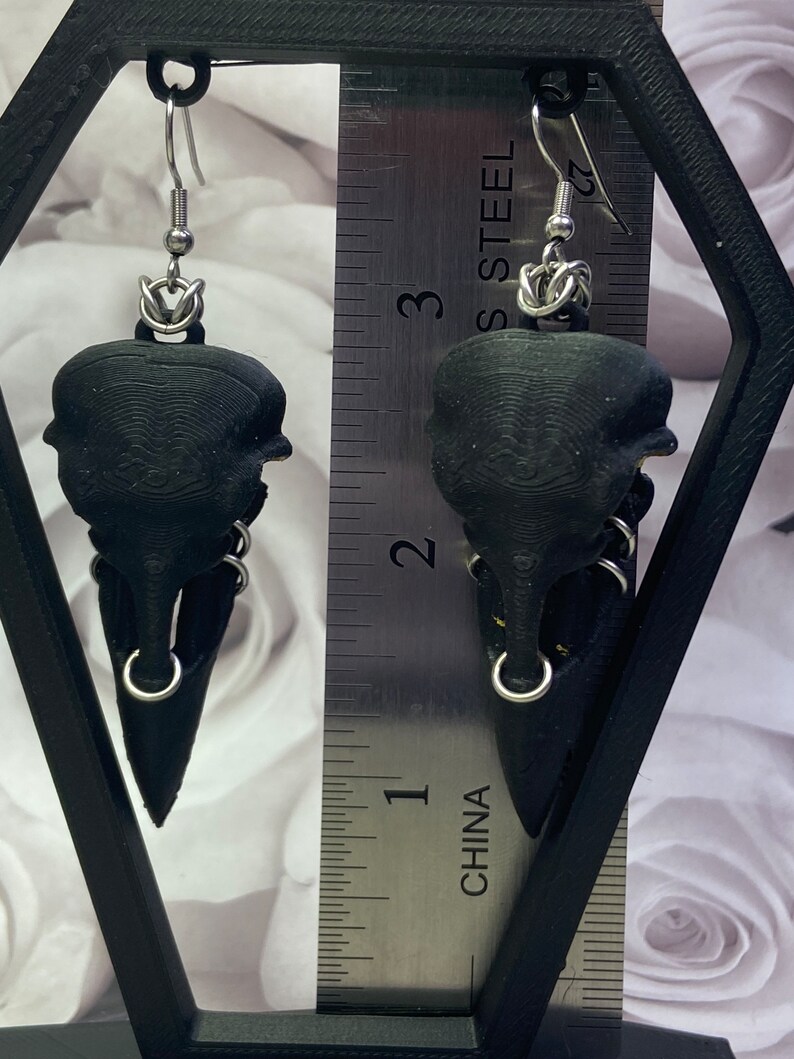 May include: A pair of black bird skull earrings with silver hoops and chains. The earrings are hanging from a ruler marked in inches and labeled "CHINA" and "STAINLESS STEEL".