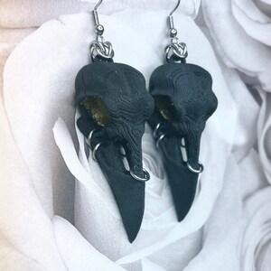 May include: A pair of black crow skull earrings with silver hoops and a gold accent. The earrings are hanging from silver hooks.
