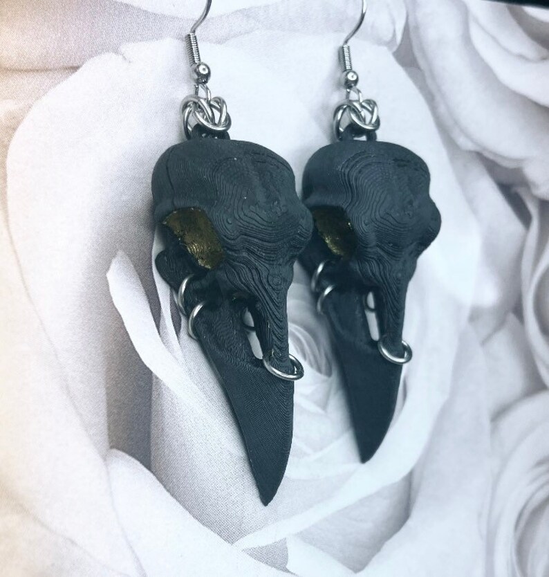May include: A pair of black crow skull earrings with silver hoops and a gold accent. The earrings are hanging from silver hooks.