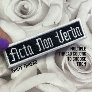 May include: A rectangular embroidered patch featuring a black background with the white text "Acta Non Verba" in a gothic-style font. The patch has a white border and includes the printed text "WHITE THREAD" and "MULTIPLE THREAD COLORS TO CHOOSE FROM".
