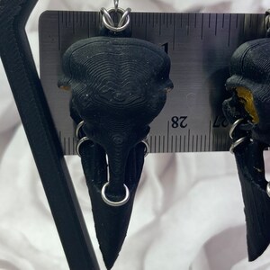 May include: A pair of black skull earrings with silver hoops. The earrings are about 1 inch long and are made of a 3D printed material.