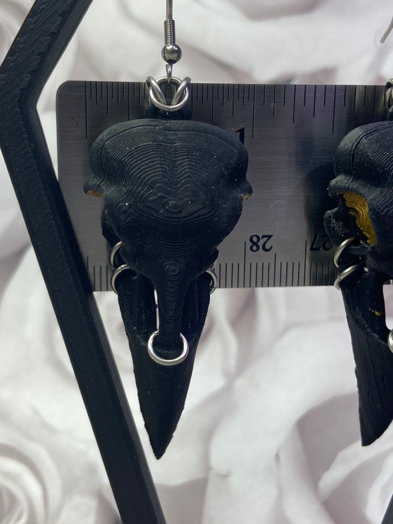 May include: A pair of black skull earrings with silver hoops. The earrings are about 1 inch long and are made of a 3D printed material.