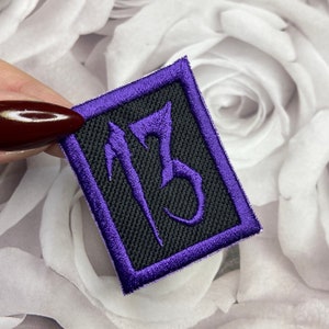 May include: Purple embroidered patch with the number 13 in a gothic font on a black background.