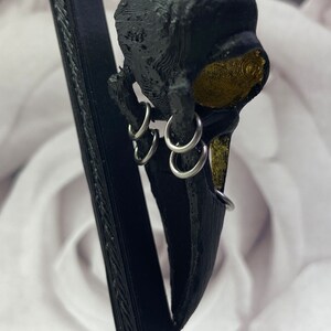 May include: Black bird skull pendant with gold accents and silver rings. The pendant is hanging from a black stand.