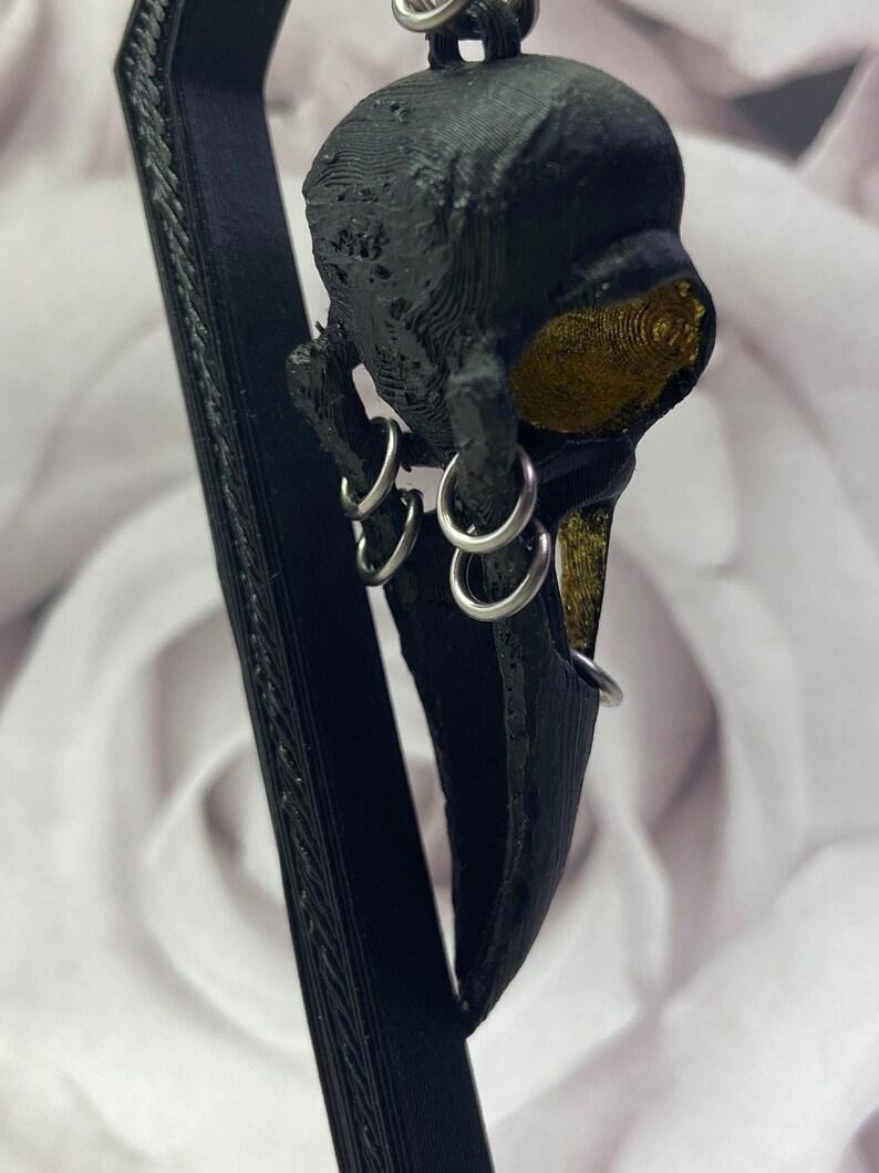 May include: Black bird skull pendant with gold accents and silver rings. The pendant is hanging from a black stand.