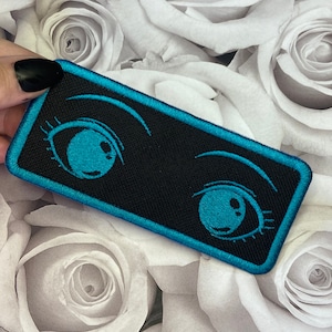 May include: A black and teal embroidered patch with two large anime eyes. The eyes are outlined in teal and have black pupils.
