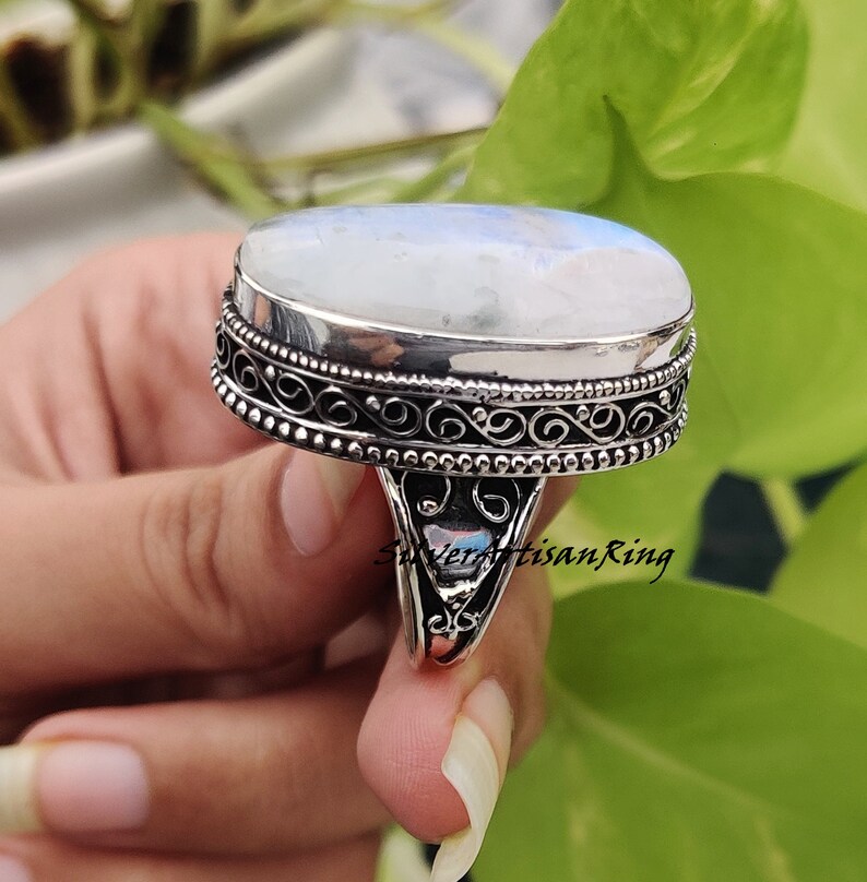 May include: A silver ring with an oval-shaped moonstone set in a filigree design. The ring has a textured band and a beaded edge.
