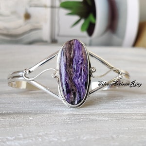 Könnte beinhalten: Ein silbernes Armband mit einem ovalen Stein in Lila und Braun. Das Armband hat ein zartes Design mit einer Spirale auf jeder Seite des Steins.