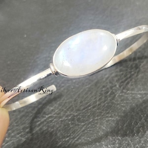 Könnte beinhalten: Ein silbernes Armreif-Armband mit einem ovalen, irisierenden Mondstein. Das Armband hat eine gehämmerten Textur. Der Text "Silver Artisan Ring" ist sichtbar. Das Armband wird von einer Hand gehalten.