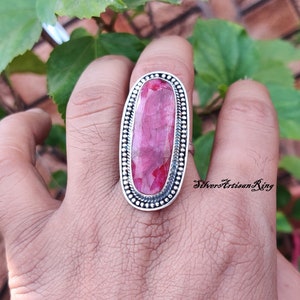 May include: A silver ring with a large, oval-shaped, pink gemstone set in a silver bezel. The ring has a beaded border around the gemstone.