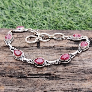 Könnte beinhalten: Ein silbernes Armband mit einem Kettenglieddesign und sechs roten Edelstein-Akzenten. Die Edelsteine sind birnenförmig und haben eine facettierte Oberfläche.