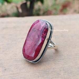 May include: A silver ring featuring a large, rectangular ruby gemstone. The ruby is a deep red hue with internal patterns. The ring has a textured edge and a simple, elegant design. The text "SilverArtisanRing" is visible.