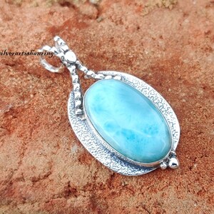 May include: A silver pendant featuring a light blue oval-shaped gemstone with a swirling pattern. The pendant is set in a silver frame with a textured design.