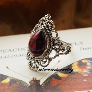 May include: A silver ring with an ornate design features a teardrop-shaped red gemstone. The ring is set against a background of a book with the word "Peacock" visible.