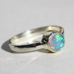 Könnte beinhalten: Ein silberner Ring mit einem runden Opal-Edelstein. Der Opal hat einen leicht bläulichen, grünen und rosafarbenen irisierenden Schimmer. Der Ring ist mit den Worten "Silver Artisan Ring" beschriftet.