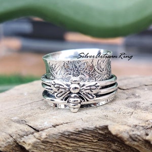 May include: A silver spinning ring with a bee design and floral pattern. The ring is made of sterling silver and has a smooth, polished finish.