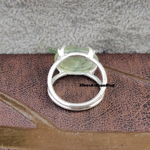 May include: A silver ring with a green gemstone set in a bezel setting. The ring is on a brown leather surface.
