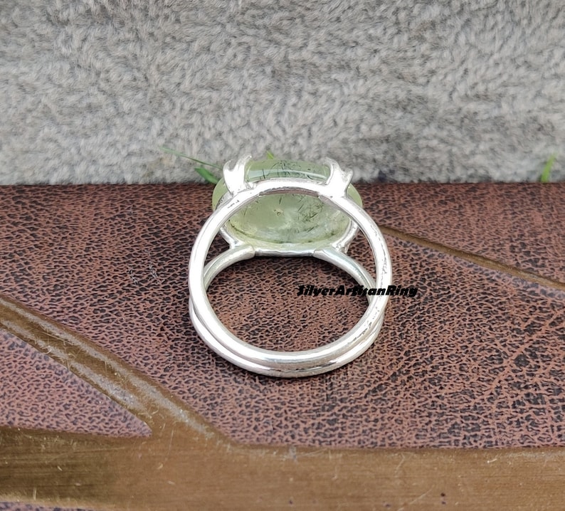 May include: A silver ring with a green gemstone set in a bezel setting. The ring is on a brown leather surface.