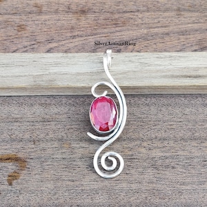 Peut inclure: Un pendentif en argent avec une pierre précieuse rouge sertie dans un motif en spirale. Le pendentif est posé sur une surface en bois.