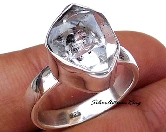 May include: A silver ring with a large, clear, faceted gemstone. The ring is marked "925" on the inside band. The text "Silver Artisan Ring" is visible on the band.