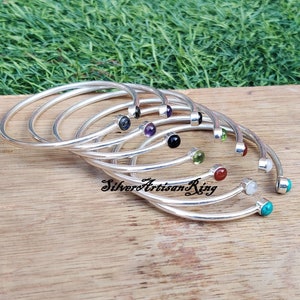 Pode incluir: Um conjunto de pulseiras de prata com pedras preciosas de diferentes cores. As pulseiras est&atilde;o empilhadas umas sobre as outras e as pedras preciosas s&atilde;o vis&iacute;veis. As pulseiras est&atilde;o sobre uma superf&iacute;cie de madeira.