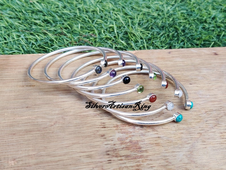 Pode incluir: Um conjunto de pulseiras de prata com pedras preciosas de diferentes cores. As pulseiras est&atilde;o empilhadas umas sobre as outras e as pedras preciosas s&atilde;o vis&iacute;veis. As pulseiras est&atilde;o sobre uma superf&iacute;cie de madeira.