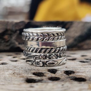May include: A stack of silver rings with various designs, including braided, smooth, and floral patterns. The rings are displayed on a rustic wooden surface, showcasing the intricate details and textures of the jewelry.