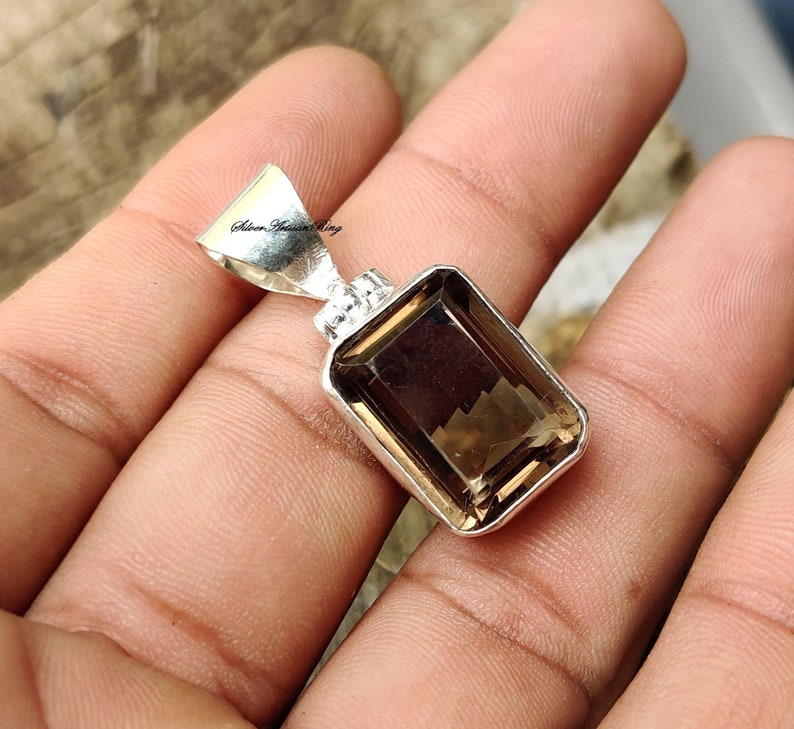Pode incluir: Um pingente de prata com uma pedra preciosa marrom. A pedra preciosa &eacute; cortada em forma retangular com bordas chanfradas. O pingente est&aacute; sobre um fundo branco.