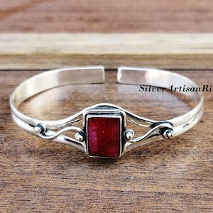 May include: A silver bangle bracelet featuring a red gemstone set in a silver frame. The bracelet has a delicate, flowing design with small silver beads on either side of the gemstone.
