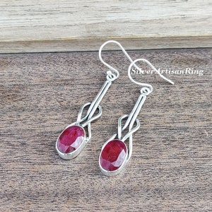 Könnte beinhalten: Ein Paar silberfarbene Ohrhänger. Jeder Ohrring hat einen ovalen, tiefroten Edelstein, der in einer silbernen Fassung gefasst ist. Das Design umfasst eine Schlaufenverzierung über dem Edelstein und einen Haken für durchbohrte Ohren.